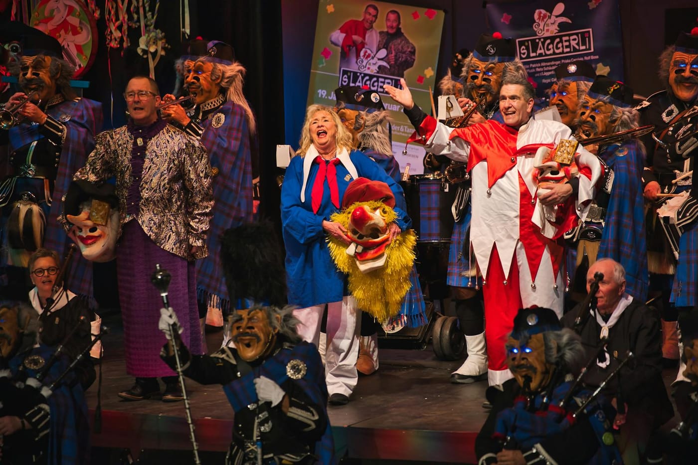 Läggerli 2025: Das Ensemble in Harlekin-Kostümen singt auf der Bühne, umringt von Guggemusikern in Larven und Kilts.